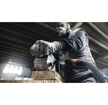 En man slipar en träbit med en slipmaskin. Han bär ett skyddsmask och skyddsglasögon.