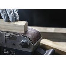 Närbild av en bandslip som slipar en träbit, manövrerad av en person med handskar.