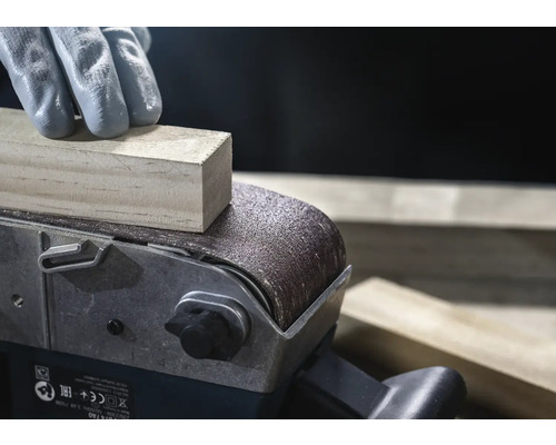 Närbild av en bandslip som slipar en träbit, manövrerad av en person med handskar.