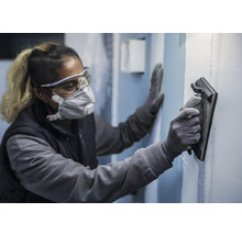 En person bär skyddsutrustning och slätar ut en vägg med ett glättningsverktyg.