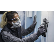 En person bär skyddsutrustning och använder en slipbräda för att slipa en vägg.