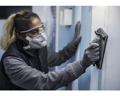 En person bär skyddsutrustning och slätar ut en vägg med ett glättningsverktyg.