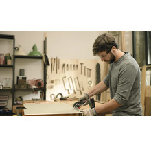 En man sågar en träplanka med en handhållen cirkelsåg. Han bär skyddsglasögon och handskar.