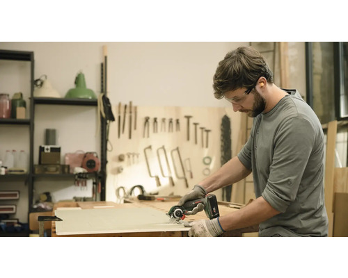 En man sågar en träplanka med en handhållen cirkelsåg. Han bär skyddsglasögon och handskar.