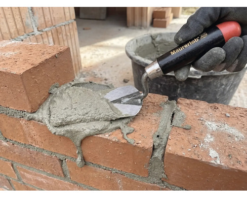 En hand med handske applicerar murbruk på en tegelvägg med en murslev