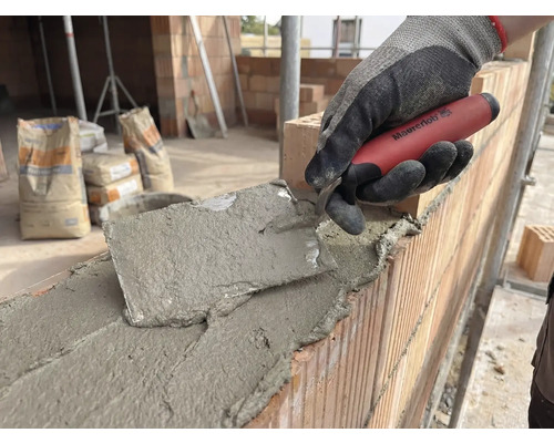 Murare arbetar med en murslev på en tegelvägg, cementsäckar i bakgrunden.