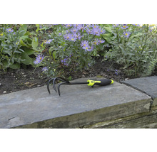 Trädgårdskultivator med tre tänder och plasthandtag framför blomsterrabatt