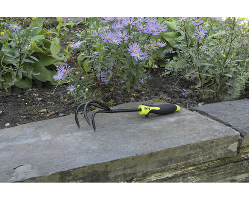 Trädgårdskultivator med tre tänder och plasthandtag framför blomsterrabatt