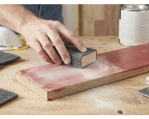 En hand slipar en lackad träskiva med ett slipvlies på en slipkloss. Slipdamm är synligt på träet.