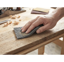 Hand som bearbetar en träplanka med slipvlies på en ljus arbetsbänk med hyvel och träspån.