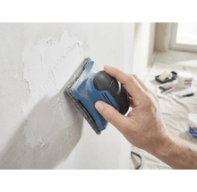 En hand slipar en spacklad vägg med en handslipmaskin och slipvlies.