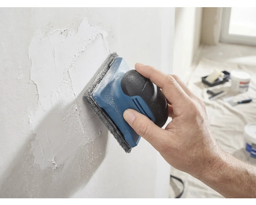 En hand slipar en spacklad vägg med en handslipmaskin och slipvlies.