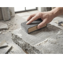 En hand för ett slipblock av trä med grå slipvlies över ytan på en grov natursten.