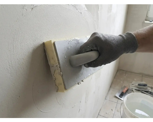 Hand med arbetshandske som använder ett svampbräde för att jämna ut puts på en vägg.