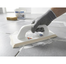 En person med skyddshandske applicerar vit fogmassa på grå golvplattor med ett epoxifogbräde.