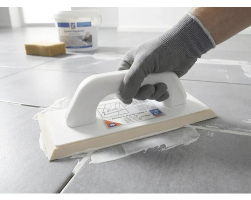 En person med skyddshandske applicerar vit fogmassa på grå golvplattor med ett epoxifogbräde.