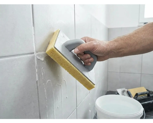 En person rengör nyligen fogat väggkakel med en svampbräda och vatten.