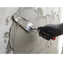 En hand med arbetshandske applicerar murbruk på en vägg med en murarslev från Maurerlob.