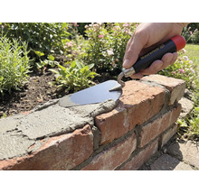 Hand med Maurerlob murarslev som applicerar bruk på en tegelmur i en trädgård.