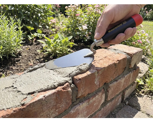 Hand med Maurerlob murarslev som applicerar bruk på en tegelmur i en trädgård.