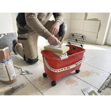 En knästående person rengör plattor med ett svampbräde och ett rött tvättset på hjul i ett ljust rum.