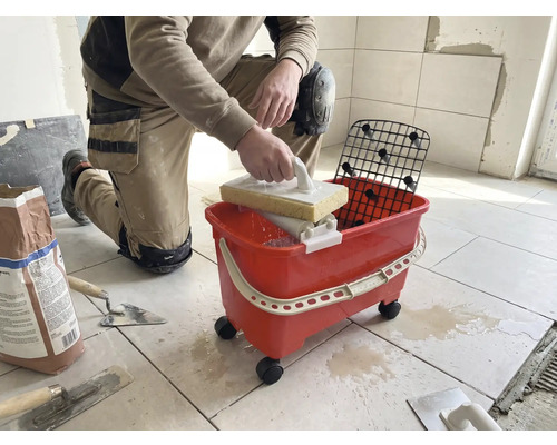 En knästående person rengör plattor med ett svampbräde och ett rött tvättset på hjul i ett ljust rum.