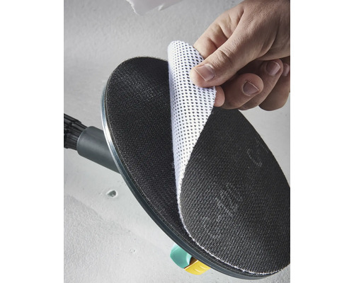 En hand fäster en vit nätsliprondell på den svarta runda slipplattan på en väggslipmaskin.