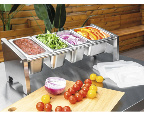 Behållare för tillbehör i rostfritt stål med fyra fack, fyllda med färska grönsaker och sås på ett arbetsbord.