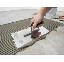 En hand applicerar fästmassa för kakel på golvet med en tandad spackel i ett räfflat mönster.