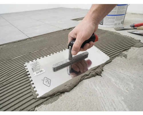 En hand applicerar fästmassa för kakel på golvet med en tandad spackel i ett räfflat mönster.