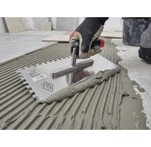 Hand med arbetshandske applicerar fästmassa för kakel på golvet med en kuggspackel av rostfritt stål.