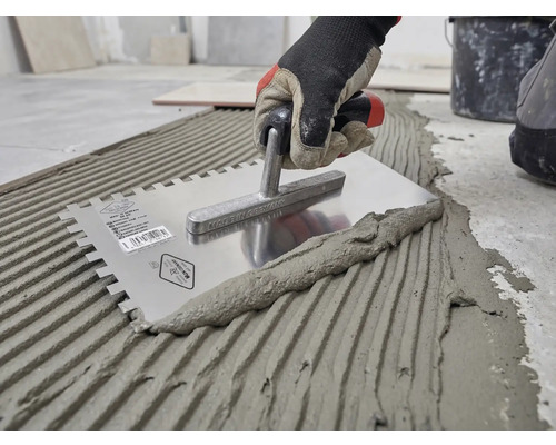 Hand med arbetshandske applicerar fästmassa för kakel på golvet med en kuggspackel av rostfritt stål.