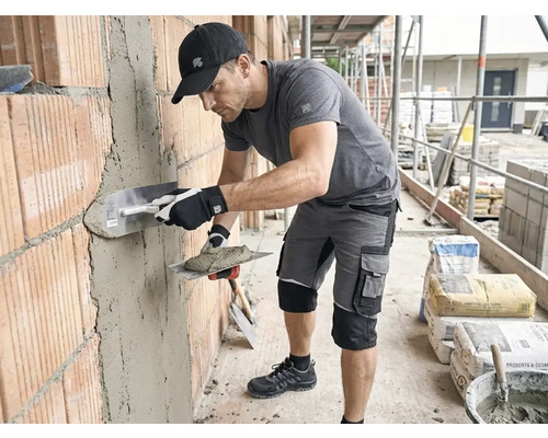 Man i arbetskläder putsar en tegelvägg på en byggarbetsplats med en putsbräda och murbruk.