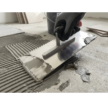 En person applicerar fästmassa på ett betonggolv med en tandspackel av metall.