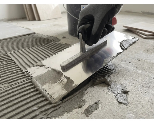 En person applicerar fästmassa på ett betonggolv med en tandspackel av metall.