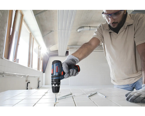 Person som använder en sladdlös slagborrmaskin från Bosch för att borra i vita kakelplattor på en byggarbetsplats.