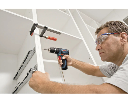 En man med skyddsglasögon monterar lådskenor i ett vitt skåp med en Bosch sladdlös borrskruvdragare och en skruvtving.