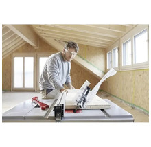 En man med skyddsglasögon kapar en träbräda med en bordscirkelsåg i ett ljust rum med träbjälkar och OSB-väggar.