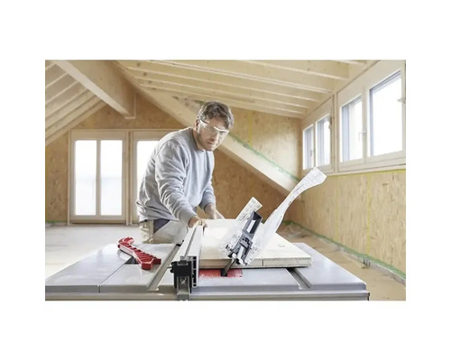 En man med skyddsglasögon kapar en träbräda med en bordscirkelsåg i ett ljust rum med träbjälkar och OSB-väggar.