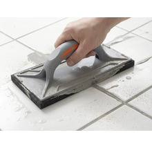 En hand för ett fogbräde över vita golvplattor för att fördela grå fogmassa.