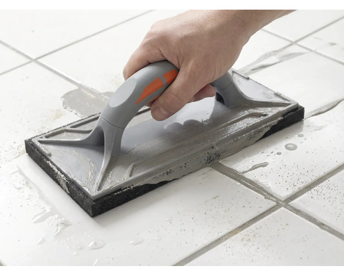 En hand för ett fogbräde över vita golvplattor för att fördela grå fogmassa.