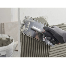 En hand med handske applicerar fästmassa jämnt på en vägg med en Akkit fixkam.