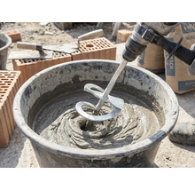 En omrörare på en borrmaskin blandar grå murbruk i en bygghink på en byggarbetsplats med tegelstenar och murbrukslevar.