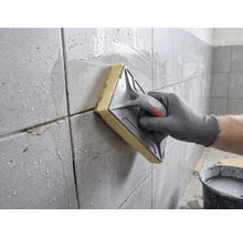 En hand rengör nyligen fogat kakel med en svampbräda och vatten.