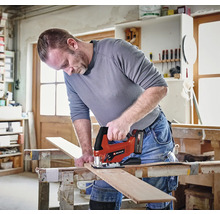 En hantverkare använder en batteridriven Einhell sticksåg för att såga en träplanka på en arbetsbänk i en ljus verkstad. Einhell Logo.