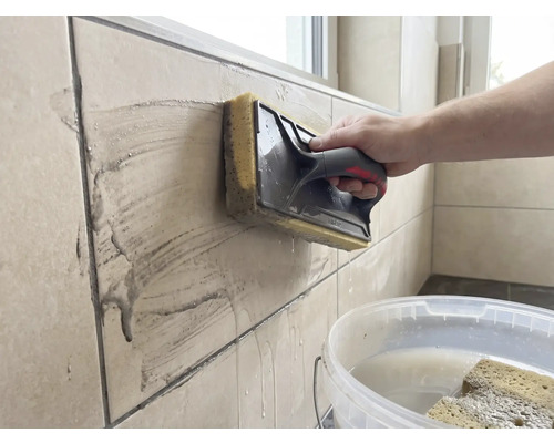 En person rengör beige väggplattor med ett svampbräda. En vit hink med vatten står i förgrunden.