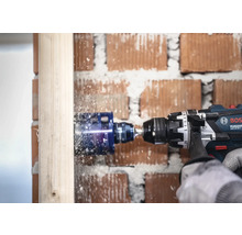 Bosch Professional sladdlös slagborrmaskin med hålsåg borrar ett hål i en träbjälke framför en tegelvägg.