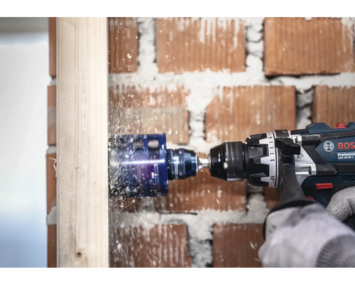 Bosch Professional sladdlös slagborrmaskin med hålsåg borrar ett hål i en träbjälke framför en tegelvägg.