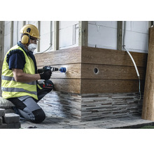 Hantverkare i skyddsutrustning borrar ett hål i en fasadpanel med trälook med hjälp av en batteridriven borrskruvdragare och en hålsåg.