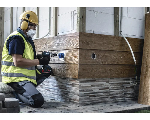 Hantverkare i skyddsutrustning borrar ett hål i en fasadpanel med trälook med hjälp av en batteridriven borrskruvdragare och en hålsåg.
